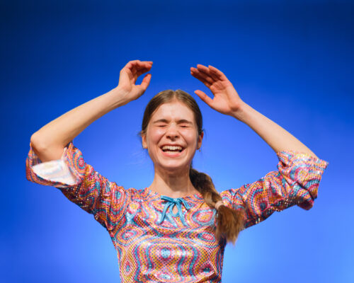 Junge Frau mit geflochtenem Zopf lacht ausgelassen mit geschlossenen Augen und erhobenen Armen vor blauem Hintergrund, sie trägt ein bunt gemustertes, glänzende
