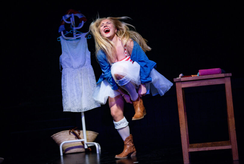 Junge Frau auf einer Theaterbühne in auffälliger Pose, mit weit geöffnetem Mund, als rufe oder schreie sie. Sie trägt ein helles Tutu, eine blau-gestreifte Ober