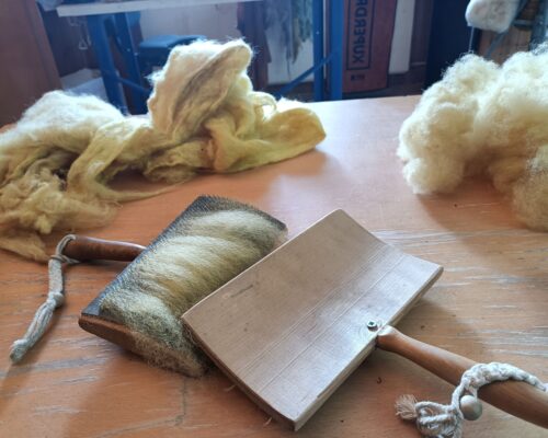 Wollkarden und rohe Wolle auf einem Arbeitstisch Auf einem Holztisch liegen gelbliche, ungekämmte Wollflocken und zwei Kardierbürsten mit Holzgriffen, auf deren Borsten ebenfalls Wolle haftet.