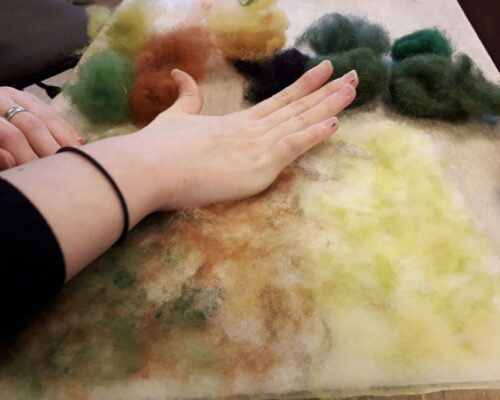 Hände beim Gestalten eines Filzbildes mit bunten Wollfasern Zwei Hände legen und streichen bunte Wollfasern in Gelb-, Grün- und Brauntönen auf einer Filzunterlage, daneben liegen weitere Faserbüschel bereit.