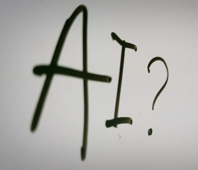 Handgeschriebener Schriftzug „AI?“ in dunkler Farbe auf hellem, unscharfem Hintergrund.