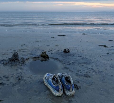 Ein Paar abgenutzte, blaue Turnschuhe steht im nassen Sand an einem ruhigen Strand. Vor den Schuhen liegt eine kleine Wasserpfütze, umgeben von dunklem Schlick