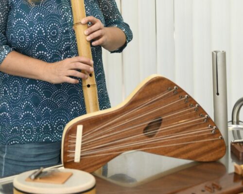 Frau spielt Flöte zwischen verschiedenen Musikinstrumenten Person in gemusterter Bluse spielt eine lange Flöte aus Holz, vor ihr stehen weitere Musikinstrumente wie ein Saiteninstrument und eine kleine Trommel auf einem