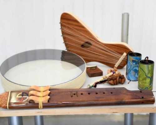 Auswahl an traditionellen Musikinstrumenten auf einem Tisch Auf einem Tisch liegen verschiedene Musikinstrumente: vorne eine längliche Holzflöte mit Verzierungen, links eine große Rahmentrommel mit hellem Fell, dahinter