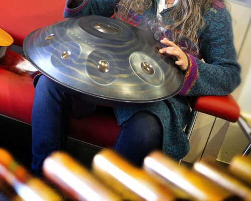 Person spielt Handpan auf rotem Sofa Person mit langen Haaren sitzt auf einem roten Sofa und hält eine metallische Handpan auf den Knien, bereit, das Instrument mit den Fingern anzuschlagen; im Hin