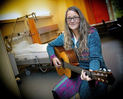 Musik im Krankenhauszimmer: Frau spielt Gitarre neben Patientenbett Eine Frau mit langen Haaren und Brille sitzt in einem Patientenzimmer neben einem Krankenhausbett und spielt auf einer Akustikgitarre. Sie trägt einen bunt gemu