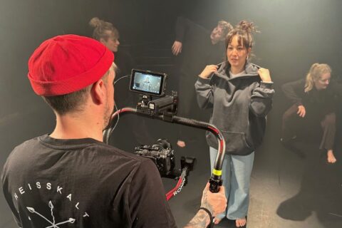 Ein Kameramann mit roter Mütze filmt eine Frau im grauen Hoodie und hellen Jeans auf einer dunklen Bühne, während im Hintergrund weitere Personen barfuß in Tanz