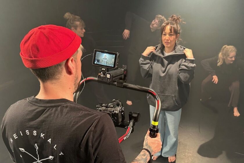 Ein Kameramann mit roter Mütze filmt eine Frau im grauen Hoodie und hellen Jeans auf einer dunklen Bühne, während im Hintergrund weitere Personen barfuß in Tanz