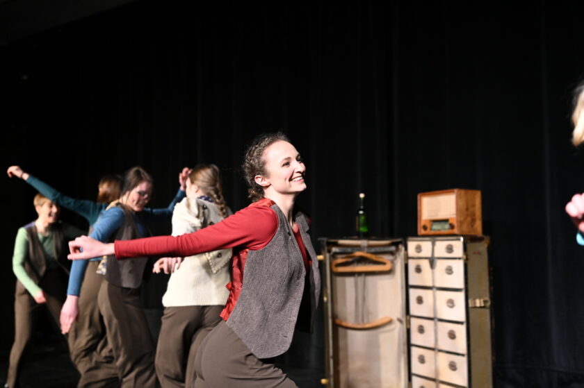 Lächelnde Tänzerin mit ausgestreckten Armen auf einer Bühne, umgeben von weiteren tanzenden Personen; im Hintergrund stehen ein alter Koffer mit Kleiderbügeln u