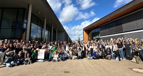 Große Gruppe junger Menschen steht und sitzt zwischen zwei modernen Hochschulgebäuden im Freien, alle lachen in die Kamera und strecken beide Arme jubelnd in di