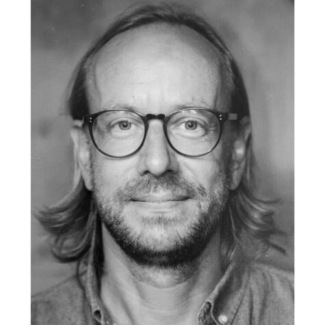 Schwarzweiß-Porträt einer mittelalten Person mit schulterlangem Haar, Brille und leichtem Bart, die direkt in die Kamera blickt und freundlich lächelt.