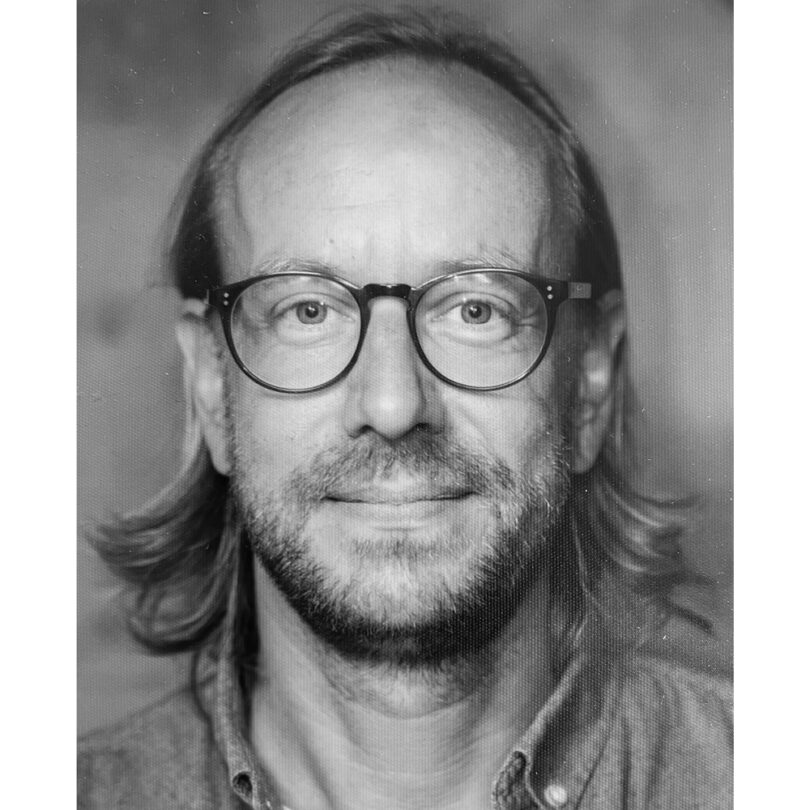 Schwarzweiß-Porträt einer mittelalten Person mit schulterlangem Haar, Brille und leichtem Bart, die direkt in die Kamera blickt und freundlich lächelt.