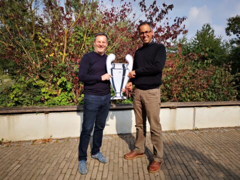 Zwei lächelnde Männer stehen im Freien vor herbstlich bunten Büschen und halten gemeinsam einen großen, gezeichneten Pokal aus Papier in die Kamera.