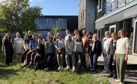 Gruppenfoto von etwa zwei Dutzend Erwachsenen, die im Freien vor einem modernen Gebäudekomplex stehen und sitzen. Die Personen tragen Namensschilder, lächeln in