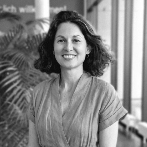 Schwarz-weiß-Porträt einer lächelnden Person mit mittellangen, lockigen Haaren und Ohrringen, die in einem hellen, modernen Innenraum vor einer Pflanze steht un