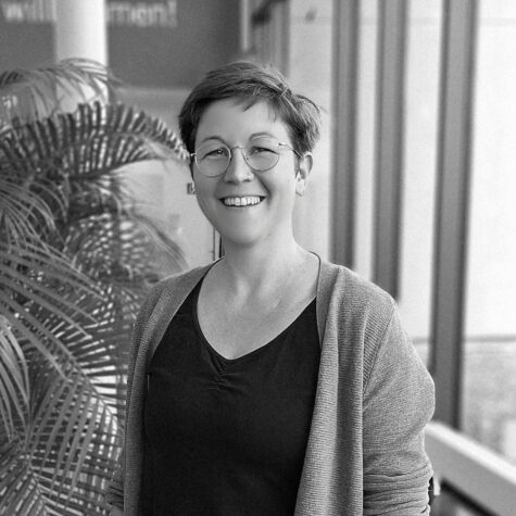 Schwarz-weiß-Porträt einer lächelnden Person mit kurz geschnittenen Haaren und runder Brille, die in einem modernen Innenraum neben einer großen Zimmerpflanze s