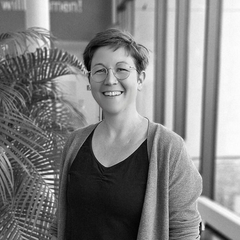 Schwarz-weiß-Porträt einer lächelnden Person mit kurz geschnittenen Haaren und runder Brille, die in einem modernen Innenraum neben einer großen Zimmerpflanze s