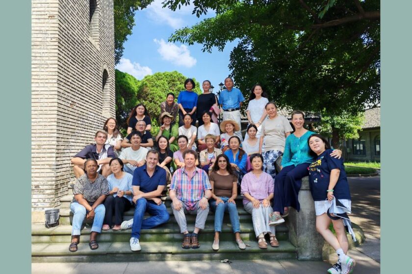 Gruppenfoto von etwa 30 Erwachsenen, die auf einer Steintreppe im Freien vor einem Backsteingebäude und unter grünen Bäumen posieren. Die Personen sitzen und st