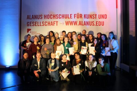 Gruppenfoto von etwa 30 lächelnden Personen vor einer beleuchteten Wand mit der Aufschrift „Alanus Hochschule für Kunst und Gesellschaft – www.alanus.edu“. Viel