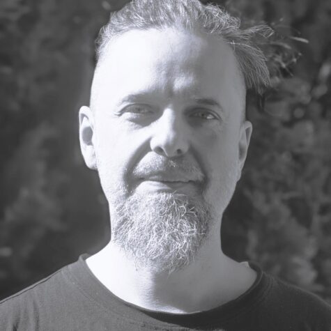 Porträtfoto eines lächelnden Erwachsenen mit kurzem Haar und längerem Kinnbart, frontal aufgenommen im Freien vor unscharfem, dunklem Hintergrund in graustufige