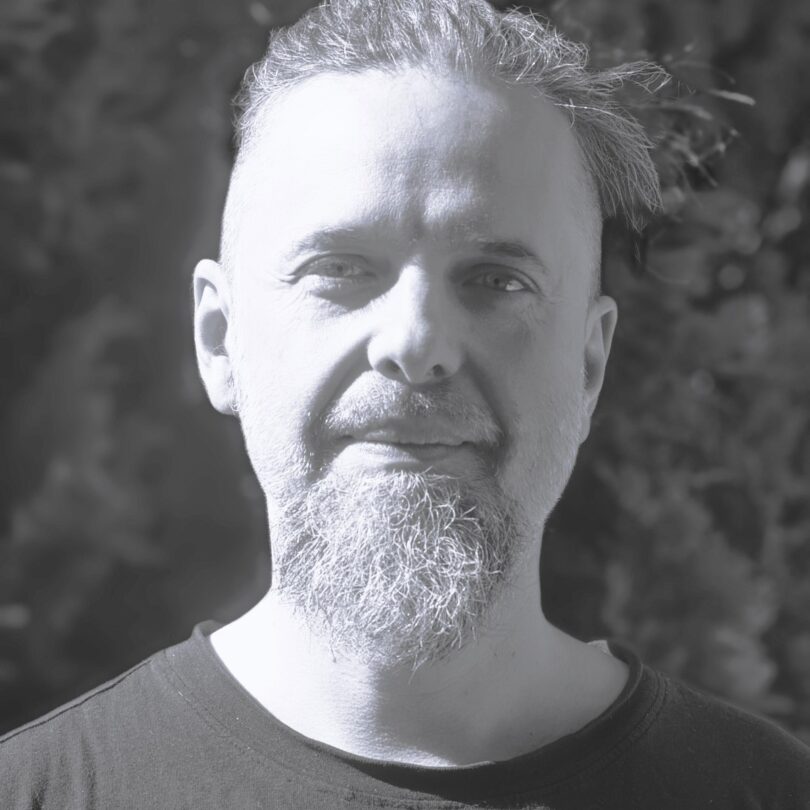 Porträtfoto eines lächelnden Erwachsenen mit kurzem Haar und längerem Kinnbart, frontal aufgenommen im Freien vor unscharfem, dunklem Hintergrund in graustufige