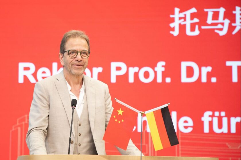 Älterer Mann im beigen Anzug spricht lächelnd an einem Rednerpult, vor ihm stehen kleine Flaggen Chinas und Deutschlands; im Hintergrund ist eine große rote Lei