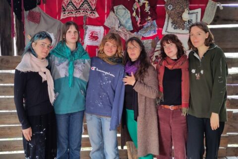 Sechs Personen stehen dicht nebeneinander vor einer Holzwand, die mit roten Tüchern und handgearbeiteten Stoffen dekoriert ist. Sie tragen lässige Kleidung mit