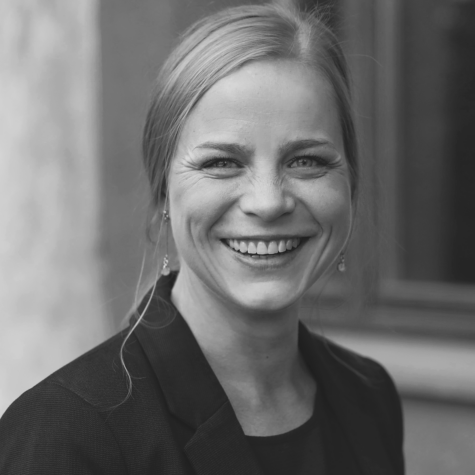 Schwarz-weiß-Porträt einer lachenden Frau mit zusammengebundenen Haaren, die eine Ohrringe und einen Blazer trägt und direkt in die Kamera blickt.