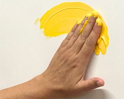 Eine Hand verstreicht mit den Fingern gelbe Farbe in einem halbkreisförmigen Strich auf einer weißen Fläche.