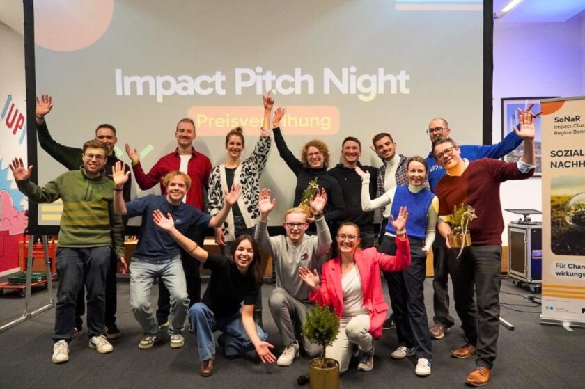 Gruppenfoto von fröhlichen Teilnehmerinnen und Teilnehmern einer Veranstaltung namens „Impact Pitch Night“. Etwa fünfzehn Menschen stehen und knien vor einer Le