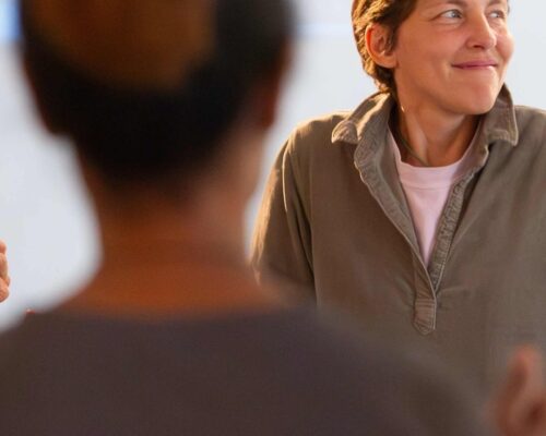 Person mit kurzem Haar und legerem Hemd lächelt zur Seite, während im unscharfen Vordergrund eine andere Person zu sehen ist, als ob sie sich in einem Gespräch