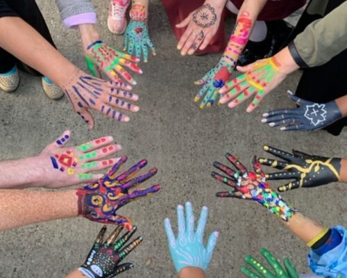 Von oben fotografierter Kreis aus ausgestreckten Händen mehrerer Personen, deren Handrücken und Finger bunt bemalt sind, mit unterschiedlichen Mustern wie Punkt