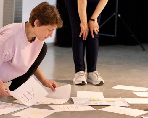 Zwei Personen in einem hellen Raum beugen sich über viele auf dem Boden verteilte Papierblätter mit Zeichnungen und handgeschriebenen Wörtern. Eine Person kniet