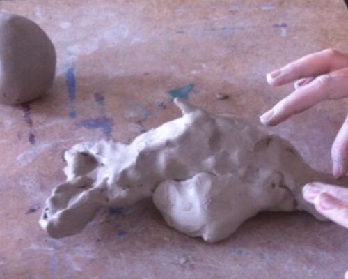 Zwei Hände formen auf einem Tisch einen unförmigen Klumpen grauen Ton, daneben liegt eine weitere kleinere Kugel aus Ton.