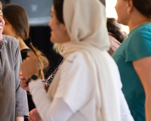 Gruppe von Menschen steht eng beieinander und unterhält sich fröhlich in einem Innenraum; im Vordergrund eine Person mit hellem Kopftuch, im Hintergrund weitere