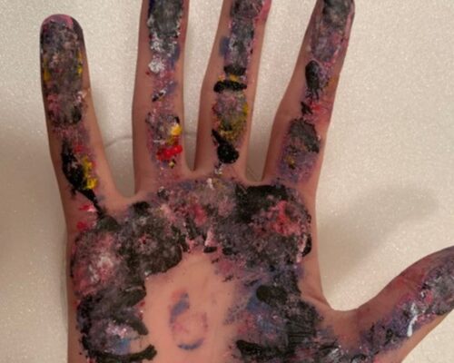 Offene Handfläche einer Person, komplett mit bunten Farbresten in Schwarz, Pink, Blau, Gelb und Weiß verschmiert, vor hellem Hintergrund.