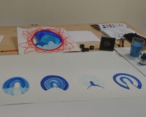 Künstlerischer Arbeitstisch mit blauen Kreis- und Bogen-Aquarellen Zeichentisch mit mehreren Blättern, auf denen in Blau- und Türkistönen halbkreisförmige und kreisförmige Aquarelle zu sehen sind. Im Hintergrund liegt ein größe