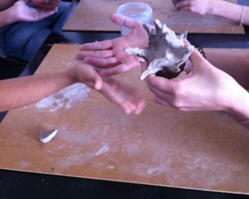 Mehrere Hände formen gemeinsam Ton auf einer Tischplatte; eine Person hält eine kleine, unregelmäßig geformte Tonskulptur, während eine andere Hand sie berührt