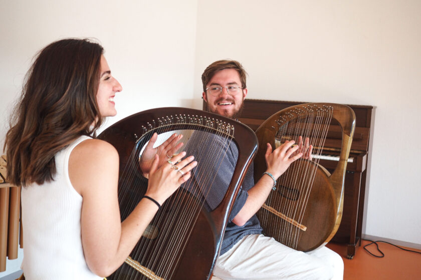 Zwei lachende Personen sitzen einander gegenüber und spielen große, bogenförmige Saiteninstrumente in einem hellen Raum, im Hintergrund steht ein Klavier.