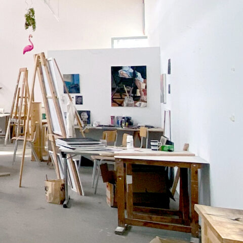 Großer, heller Kunstatelierraum mit Staffeleien, Holztischen, Stühlen und verstreuten Malutensilien. An der Rückwand hängen mehrere Gemälde, auf den Tischen ste