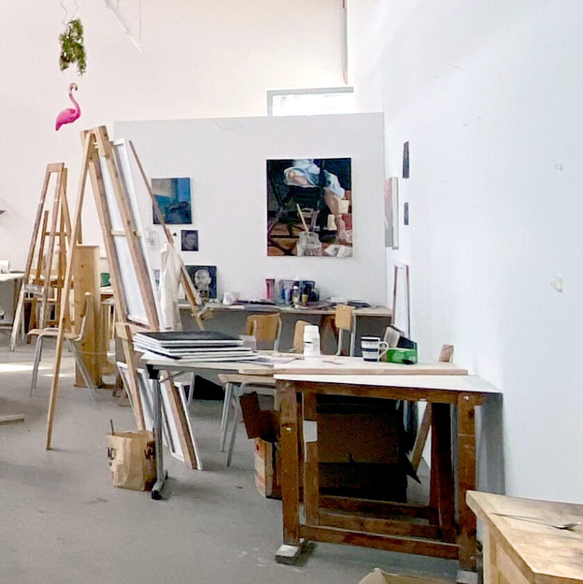 Großer, heller Kunstatelierraum mit Staffeleien, Holztischen, Stühlen und verstreuten Malutensilien. An der Rückwand hängen mehrere Gemälde, auf den Tischen ste