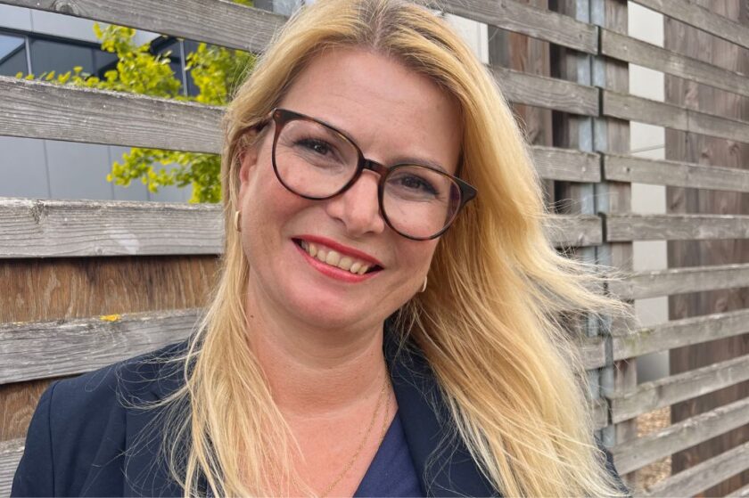 Porträt einer lächelnden Frau mit langen blonden Haaren und großer Brille, die vor einem hölzernen Lattenzaun im Freien steht.