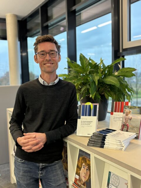 Ein lächelnder Mann mit Brille und dunklem Pullover steht in einer modernen, hellen Bibliotheks- oder Büroumgebung neben einem Regal mit mehreren aufgestellten