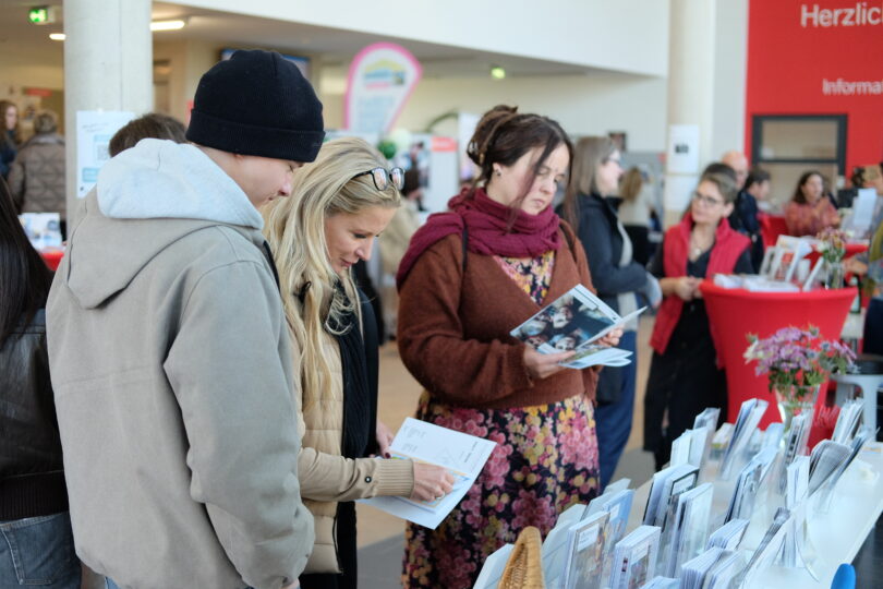 Menschen stehen in einer hellen Eingangshalle an einem Infostand und blättern in Broschüren; im Vordergrund ein junges Paar und eine Frau mit buntem Kleid, im H