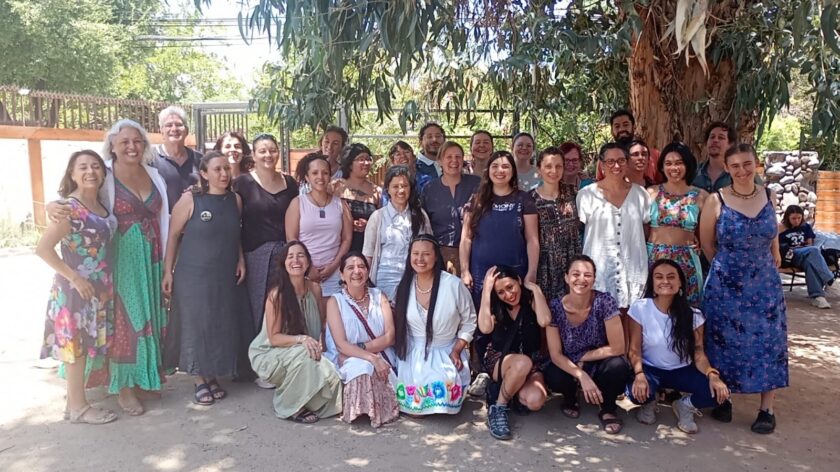 Große, bunt gekleidete Gruppe von etwa 30 lächelnden Erwachsenen posiert im Freien unter einem schattigen Baum für ein Gruppenfoto. Einige Personen knien oder s
