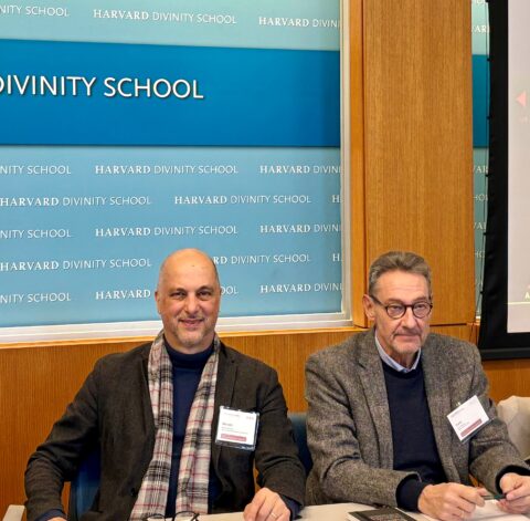 Zwei Männer sitzen nebeneinander an einem Tisch vor einer Wand mit der Aufschrift „Harvard Divinity School“. Beide tragen Jacketts und Namensschilder; der Mann