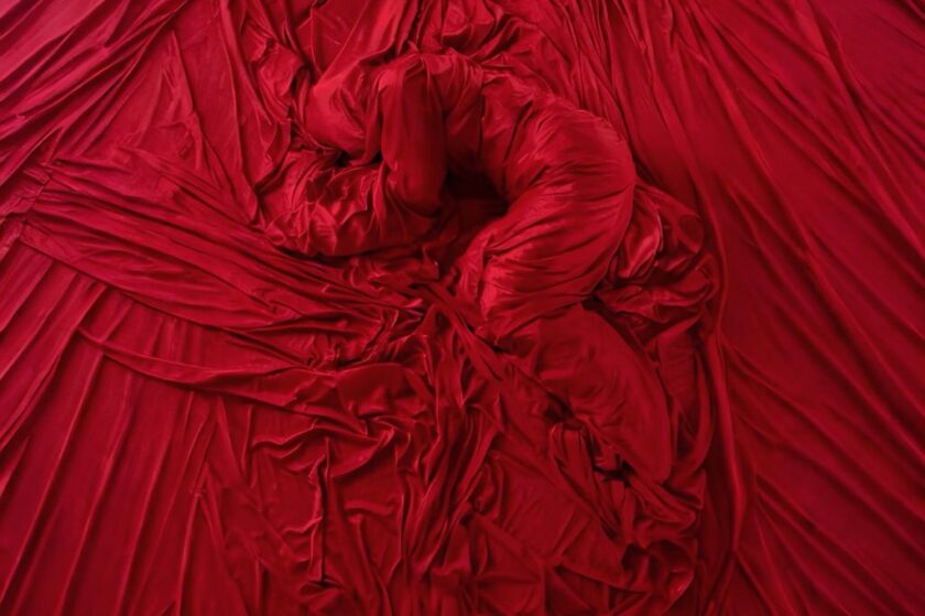 Nahaufnahme von reichlich drapiertem, knitterigem roten Stoff, der in der Mitte eine geschlungene, wurmförmige Struktur bildet.