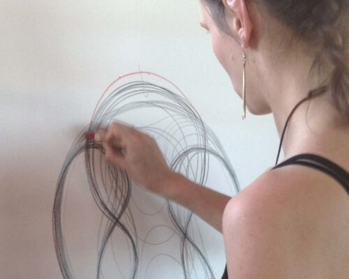 Frau beim Zeichnen dynamischer Linien auf großem Papier Eine Person zeichnet mit farbigen Stiften schwungvolle, sich überlagernde Kreise und Kurven auf ein großes weißes Blatt Papier, gesehen von hinten über die Schu