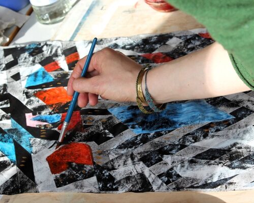 Hand beim Gestalten eines abstrakten Gemäldes Eine Person bemalt mit einem Pinsel ein abstraktes Bild mit schwarzen, weißen, roten und blauen Formen auf Papier. Der Arm mit Armbändern ist im Vordergrund zu