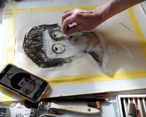 Porträtzeichnung nach Fotovorlage im Atelier Eine Hand arbeitet an einer Porträtzeichnung auf Papier, das mit gelbem Klebeband am Rand fixiert ist. Auf dem Blatt entsteht mit Kohle und Pastell das Gesicht